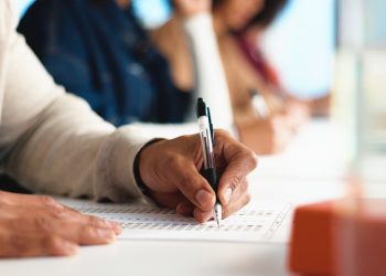 Person writing answers on a multiple choice test sheet with others blurred in the background.