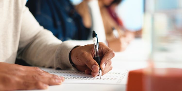 Person writing answers on a multiple choice test sheet with others blurred in the background.