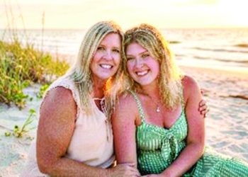 Two women smiling together on a beach at sunset with ocean waves and tall grass in the background.