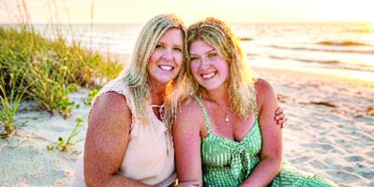 Two women smiling together on a beach at sunset with ocean waves and tall grass in the background.