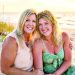 Two women smiling together on a beach at sunset with ocean waves and tall grass in the background.