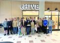 A group of students pose indoors in front of an eHIVE sign holding project displays from a school design activity.