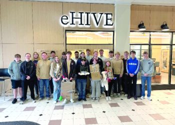 A group of students pose indoors in front of an eHIVE sign holding project displays from a school design activity.
