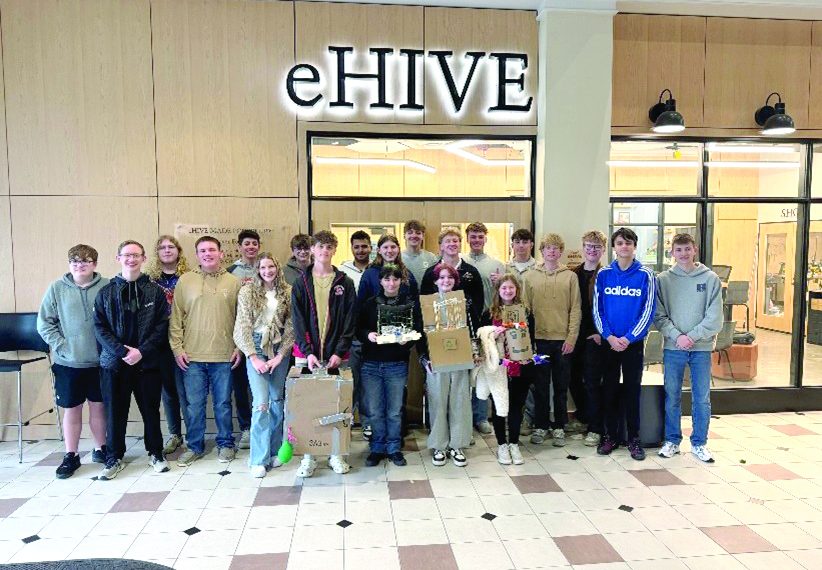 A group of students pose indoors in front of an eHIVE sign holding project displays from a school design activity.