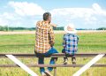 A man and child sit on a fence overlooking a farm field as the man points into the distance.
