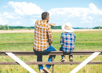 A man and child sit on a fence overlooking a farm field as the man points into the distance.
