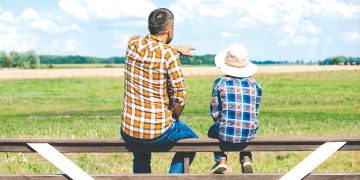 A man and child sit on a fence overlooking a farm field as the man points into the distance.
