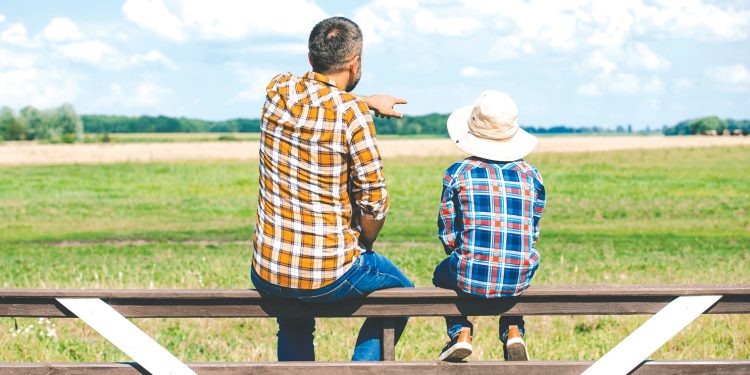 A man and child sit on a fence overlooking a farm field as the man points into the distance.