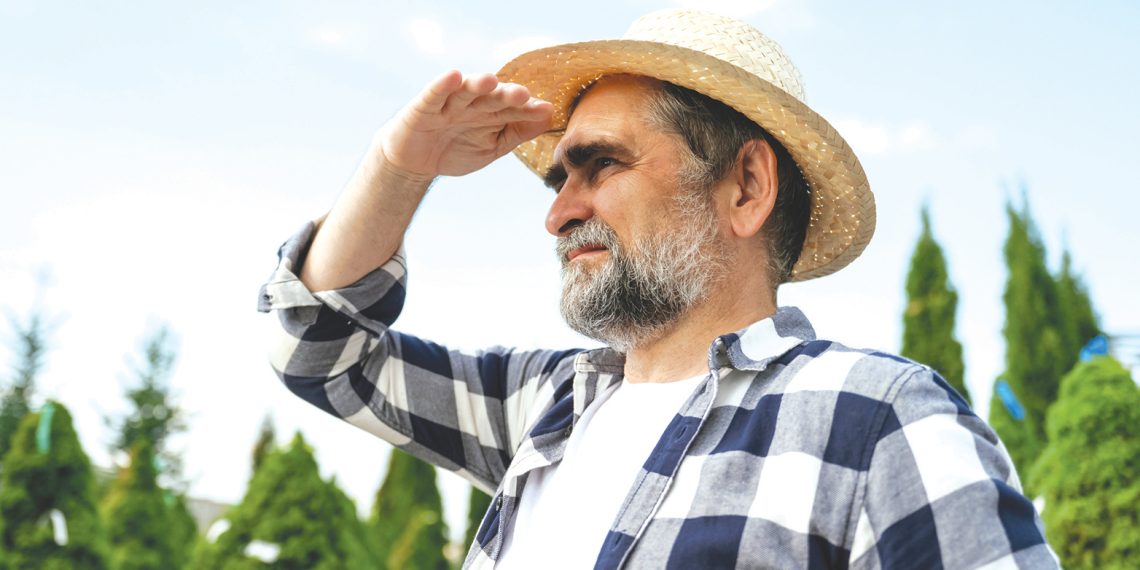 Man in a straw hat shades his eyes while looking into the distance outdoors.