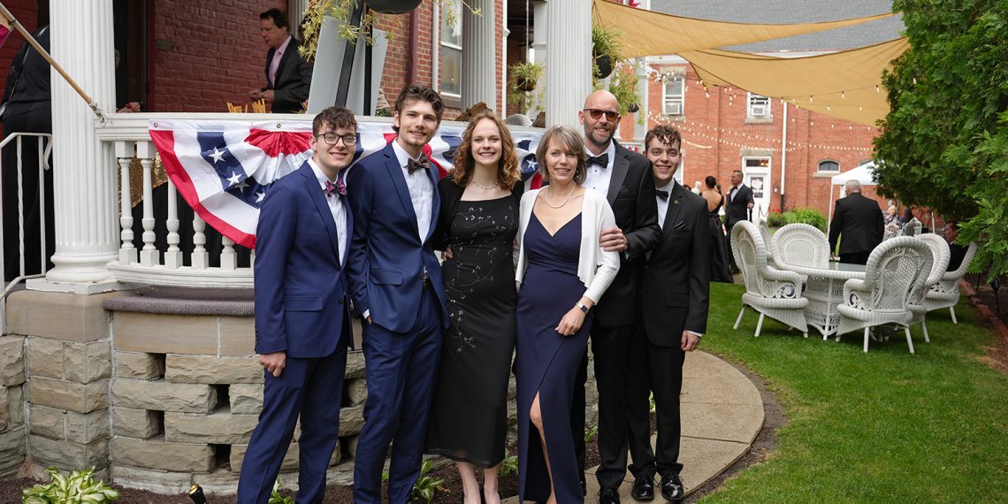 Six people dressed in formal wear pose together outside a decorated brick building during an outdoor event.