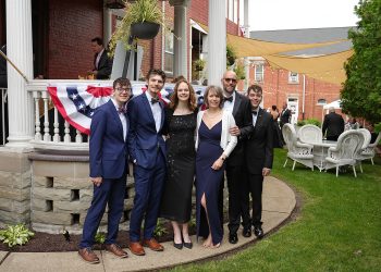 Six people dressed in formal wear pose together outside a decorated brick building during an outdoor event.