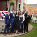 Six people dressed in formal wear pose together outside a decorated brick building during an outdoor event.