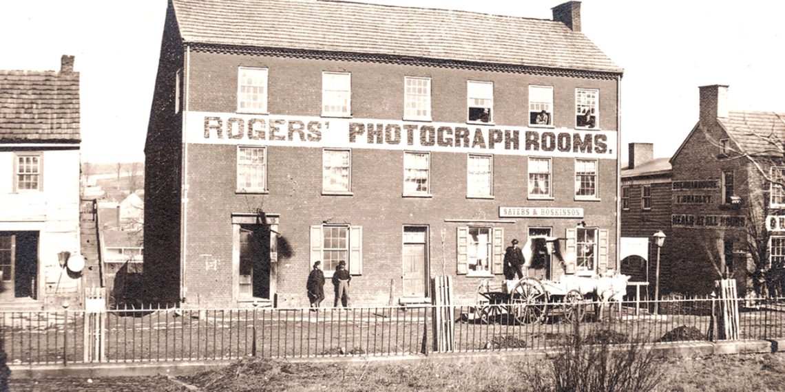 A historic black and white photo shows the Rogers’ Photograph Rooms building with people and a horse-drawn wagon outside.