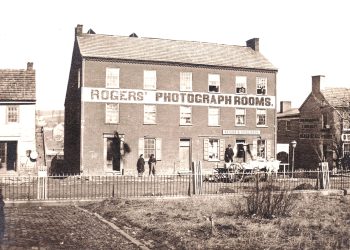 A historic black and white photo shows the Rogers’ Photograph Rooms building with people and a horse-drawn wagon outside.