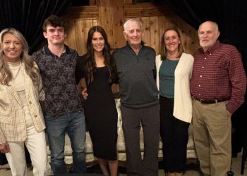 Six people pose together for a group photo inside a rustic indoor setting with wooden walls.