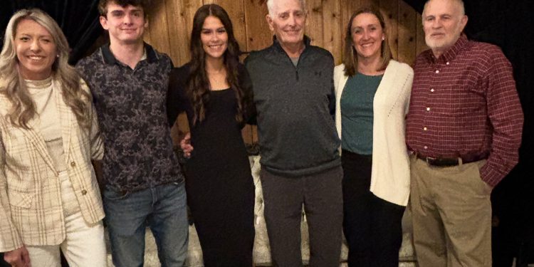 Six people pose together for a group photo inside a rustic indoor setting with wooden walls.