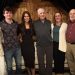 Six people pose together for a group photo inside a rustic indoor setting with wooden walls.