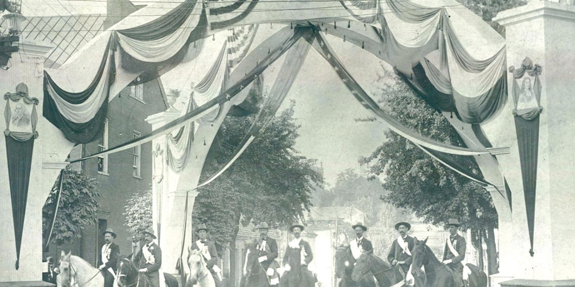 A vintage black and white photo shows riders on horseback gathered beneath a patriotic arch decorated with American flags and banners.