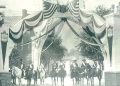 A vintage black and white photo shows riders on horseback gathered beneath a patriotic arch decorated with American flags and banners.