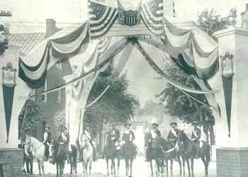 A vintage black and white photo shows riders on horseback gathered beneath a patriotic arch decorated with American flags and banners.