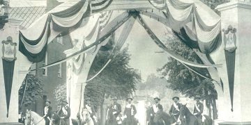 A vintage black and white photo shows riders on horseback gathered beneath a patriotic arch decorated with American flags and banners.