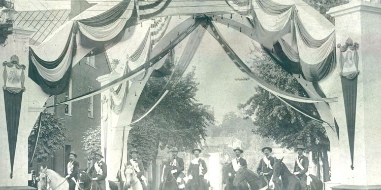 A vintage black and white photo shows riders on horseback gathered beneath a patriotic arch decorated with American flags and banners.
