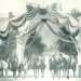 A vintage black and white photo shows riders on horseback gathered beneath a patriotic arch decorated with American flags and banners.