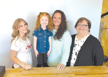 Four women and girls smile together behind a wooden counter inside a bright business setting.