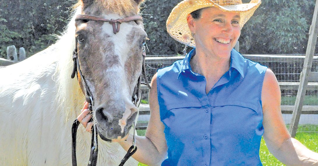 A woman in western attire stands beside a horse in a sunny outdoor pasture setting.