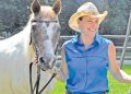 A woman in western attire stands beside a horse in a sunny outdoor pasture setting.