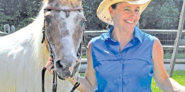 A woman in western attire stands beside a horse in a sunny outdoor pasture setting.