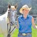 A woman in western attire stands beside a horse in a sunny outdoor pasture setting.