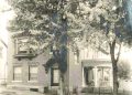 A vintage sepia photograph shows a large brick house with a covered porch and trees lining the street.