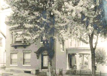 A vintage sepia photograph shows a large brick house with a covered porch and trees lining the street.
