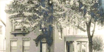 A vintage sepia photograph shows a large brick house with a covered porch and trees lining the street.