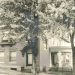 A vintage sepia photograph shows a large brick house with a covered porch and trees lining the street.