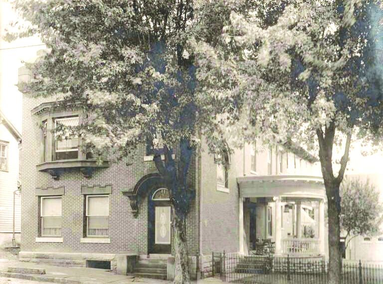 A vintage sepia photograph shows a large brick house with a covered porch and trees lining the street.