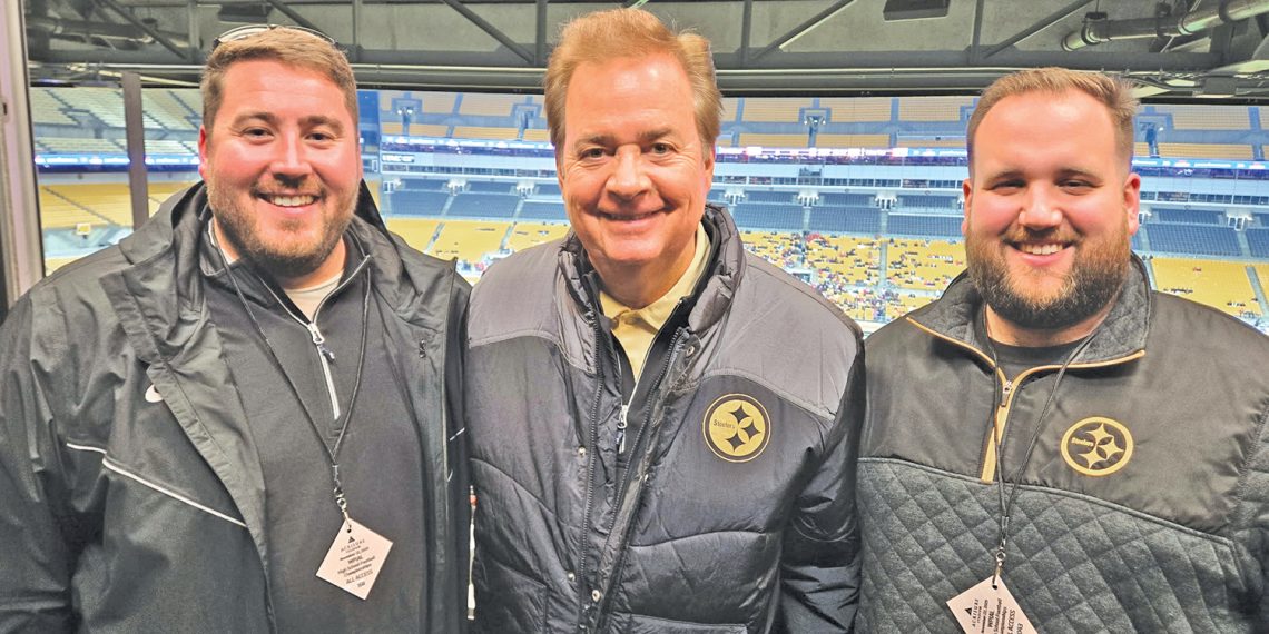Three men wearing Pittsburgh Steelers apparel pose together inside a stadium media area overlooking the field.