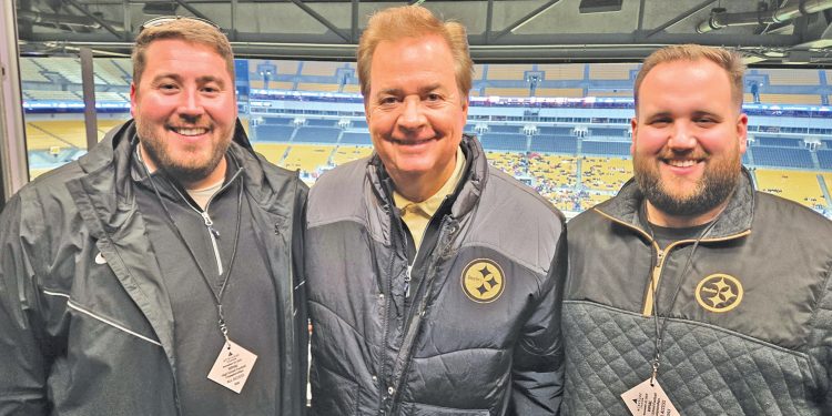 Three men wearing Pittsburgh Steelers apparel pose together inside a stadium media area overlooking the field.