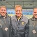 Three men wearing Pittsburgh Steelers apparel pose together inside a stadium media area overlooking the field.