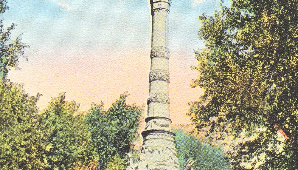 A tall stone monument with a statue on top stands in a tree-lined park under a blue sky.