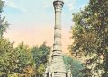 A tall stone monument with a statue on top stands in a tree-lined park under a blue sky.