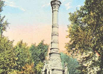 A tall stone monument with a statue on top stands in a tree-lined park under a blue sky.