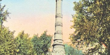 A tall stone monument with a statue on top stands in a tree-lined park under a blue sky.
