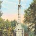 A tall stone monument with a statue on top stands in a tree-lined park under a blue sky.