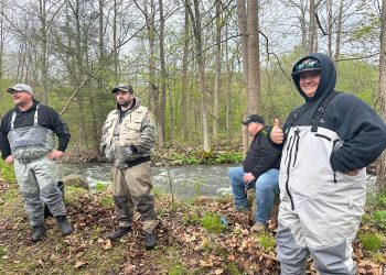 Four men in fishing waders stand beside a wooded creek during a fishing trip.