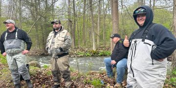 Four men in fishing waders stand beside a wooded creek during a fishing trip.