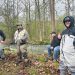 Four men in fishing waders stand beside a wooded creek during a fishing trip.