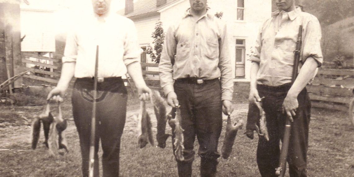 A vintage black and white photo shows three young men with rifles and game birds standing outside on a farm.
