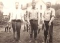 A vintage black and white photo shows three young men with rifles and game birds standing outside on a farm.